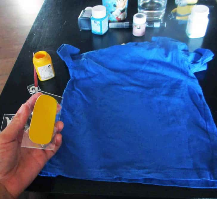 A hand holds a craft foam stamp painted with yellow paint over a blue t-shirt.