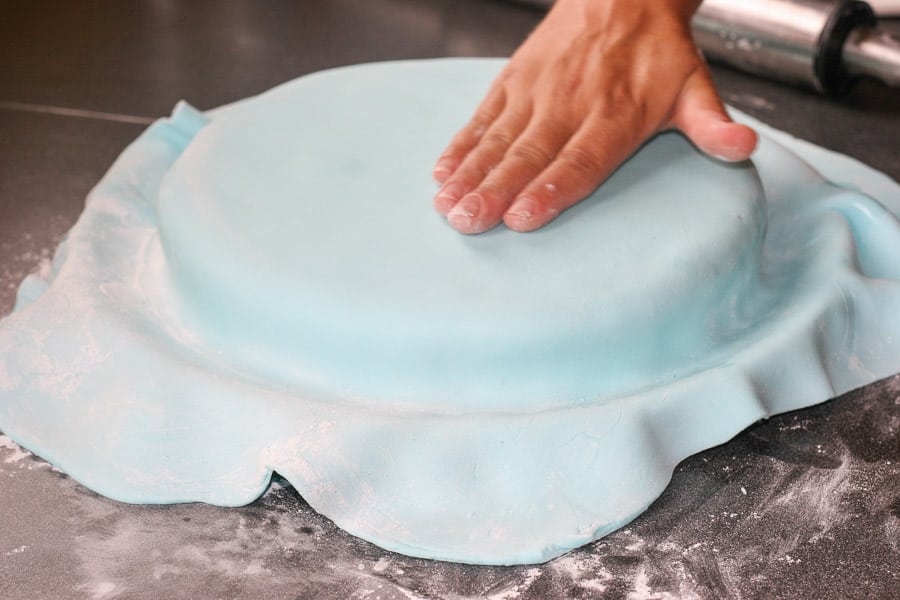 A hand smoothing blue fondant over a round cake. 