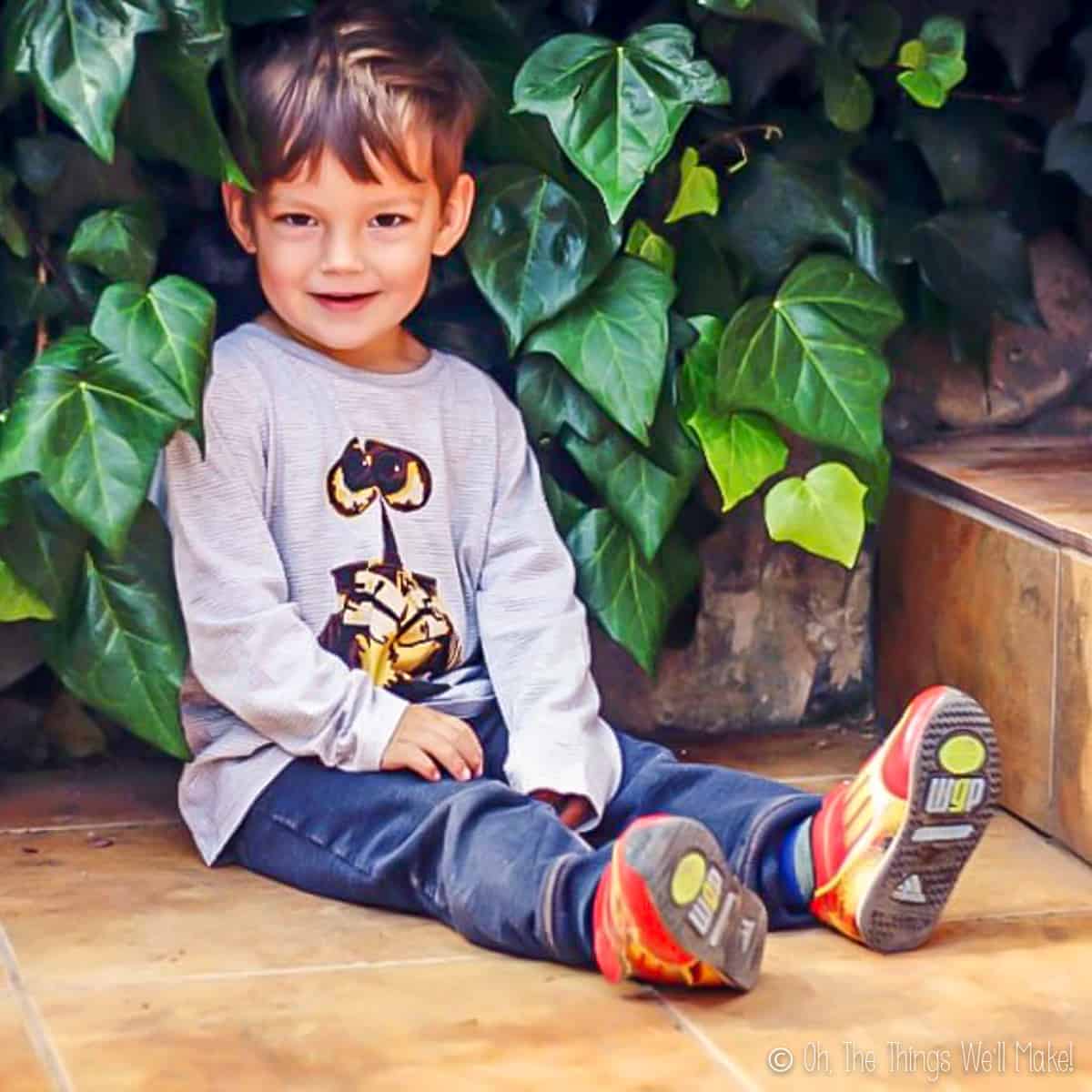 A young boy sitting on the ground with plants behind, wearing a grey wall-E t-shirt