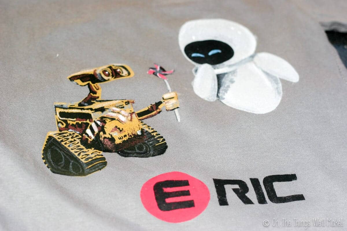 Closeup of a finished t-shirt with painted Wall-E and Eve and the name Eric painted in black with the E in a red circle, similar to the Wall-E font.