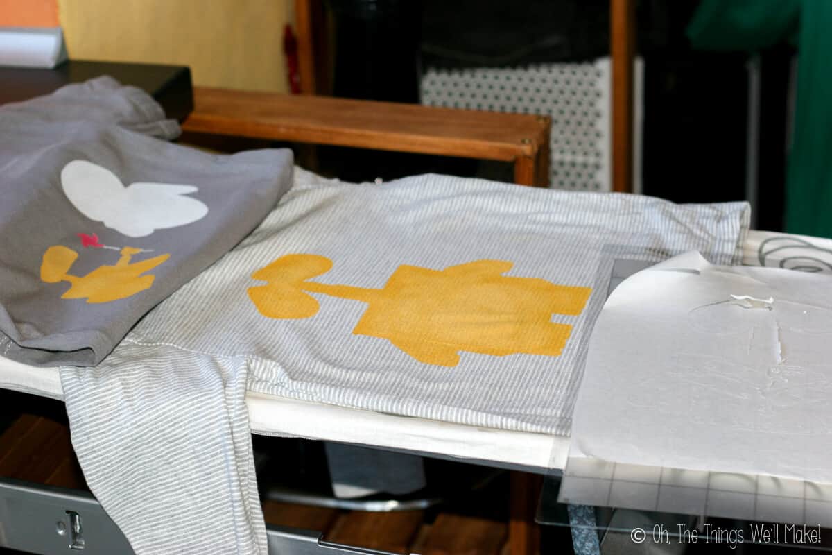 Two grey shirts placed on an ironing board. One grey shirt has yellow painted Wall-E, white painted Eve, and red pinwheel. Other shirt has a large yellow painted Wall-E.