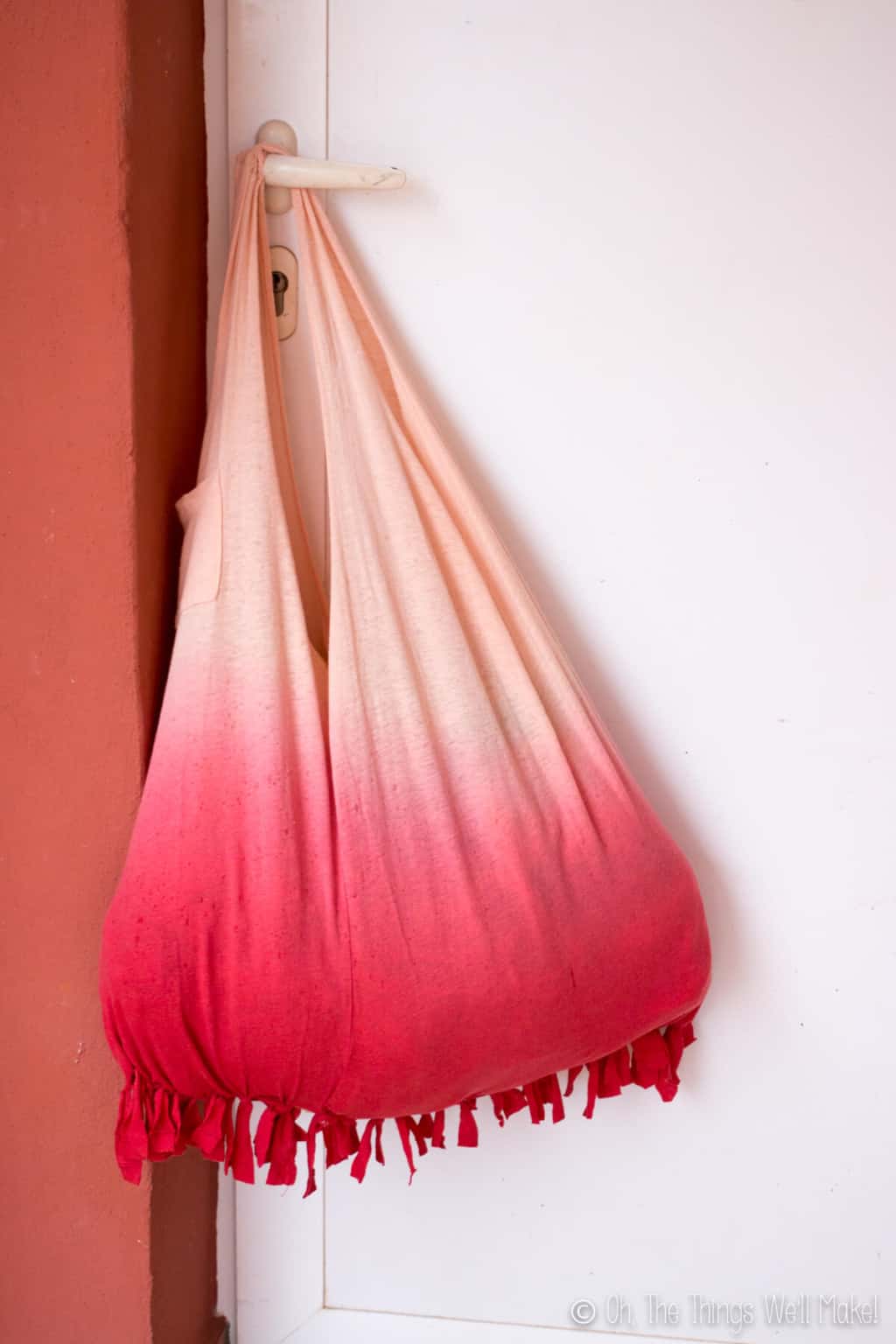 An ombre peach red no sew fringed bottom t-shirt bag hanging on a door knob of a white door.