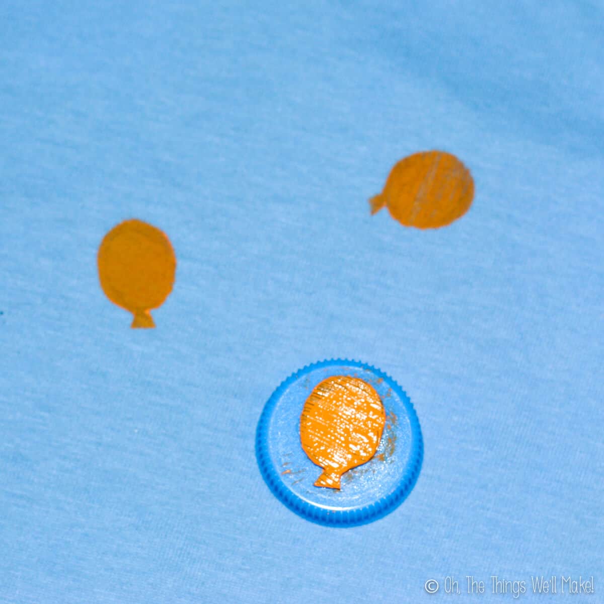 Close up of homemade balloon stamp made from an old plastic bottle cap and craft foam cut up into a balloon. Beside the stamp is two painted yellow stamp marks on a blue fabric.