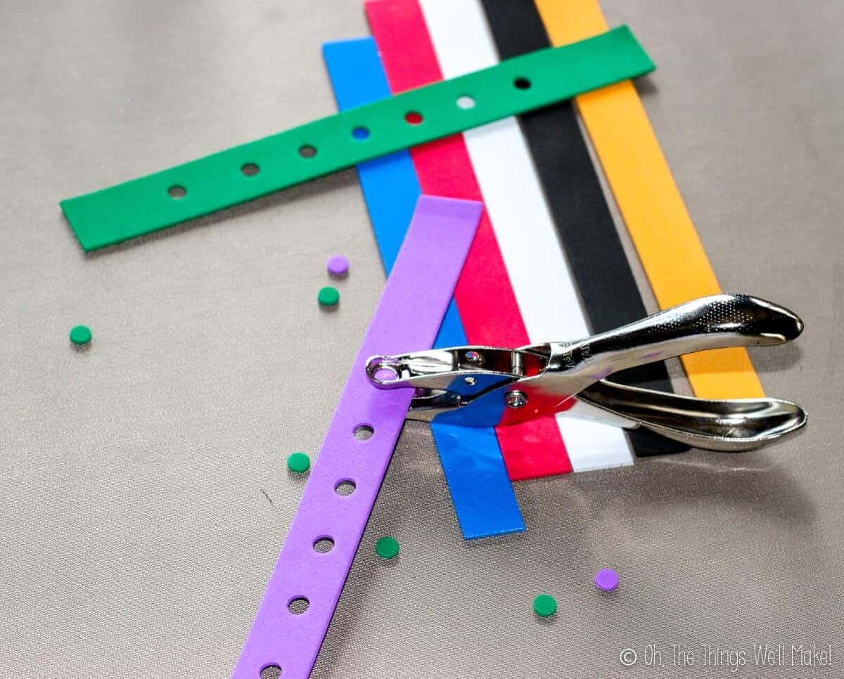 Close up of several strips of colorful craft foam with one green strip already lined with holes and a purple strip that has some holes and a hole puncher on it.