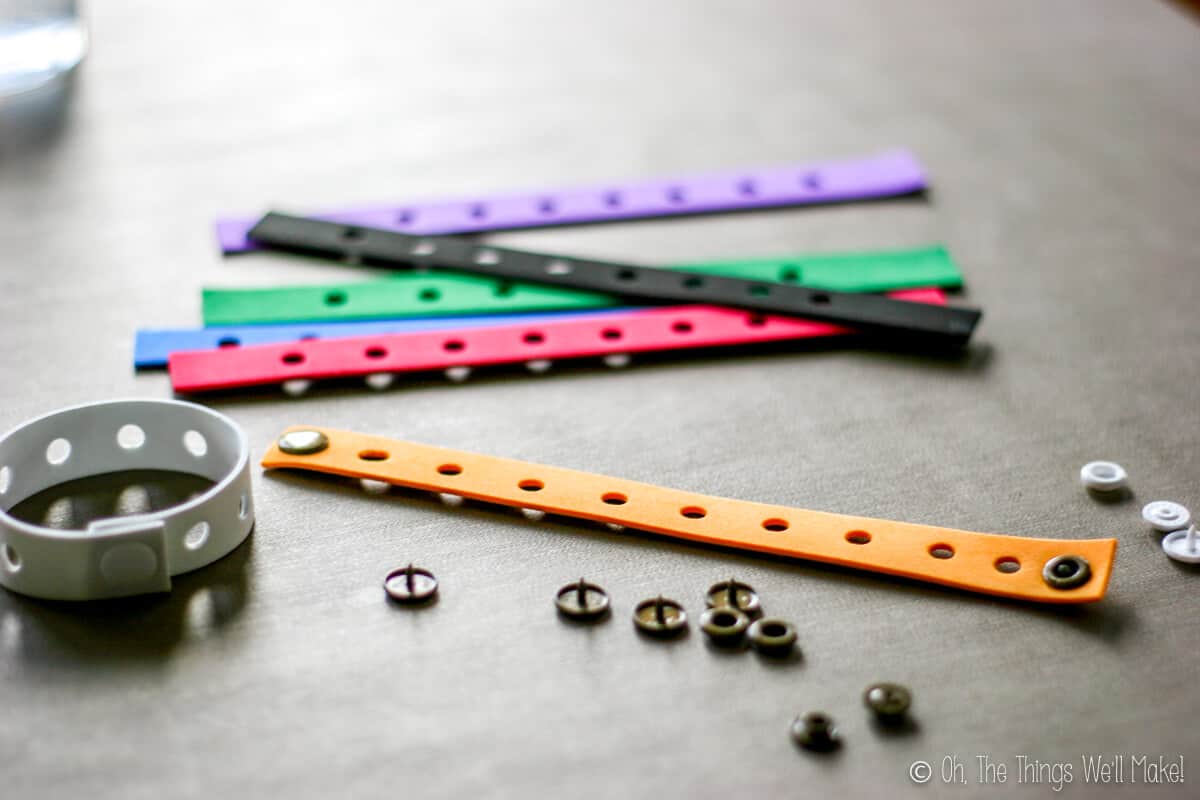 Close up of craft foam strips with holes in all of them with one white bracelet closed and one orange strip laid flat with plastic snaps already attached. There are several plastic snaps scattered around the strips.