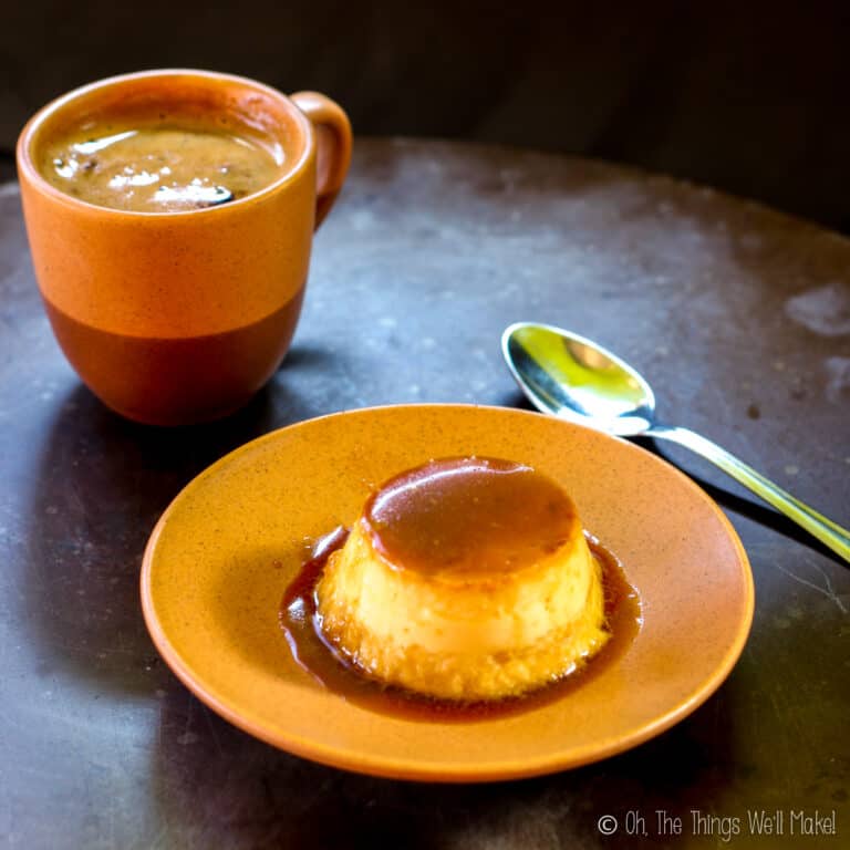 A cup of coffee and a plate with coconut flan dessert sit on a brown table, with a spoon placed beside them.