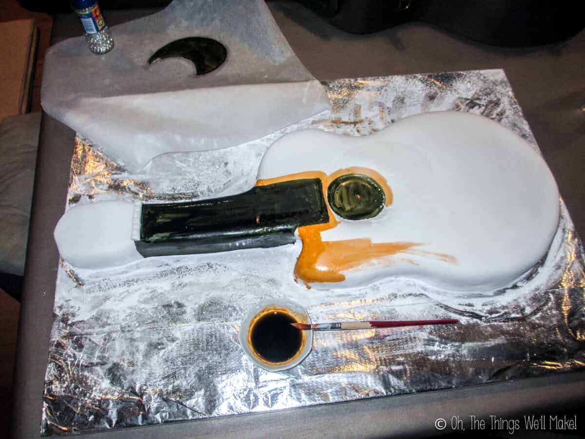 Top view of a partially painted fondant guitar cake in black and orange, with a bowl of food coloring and a paintbrush by its side.