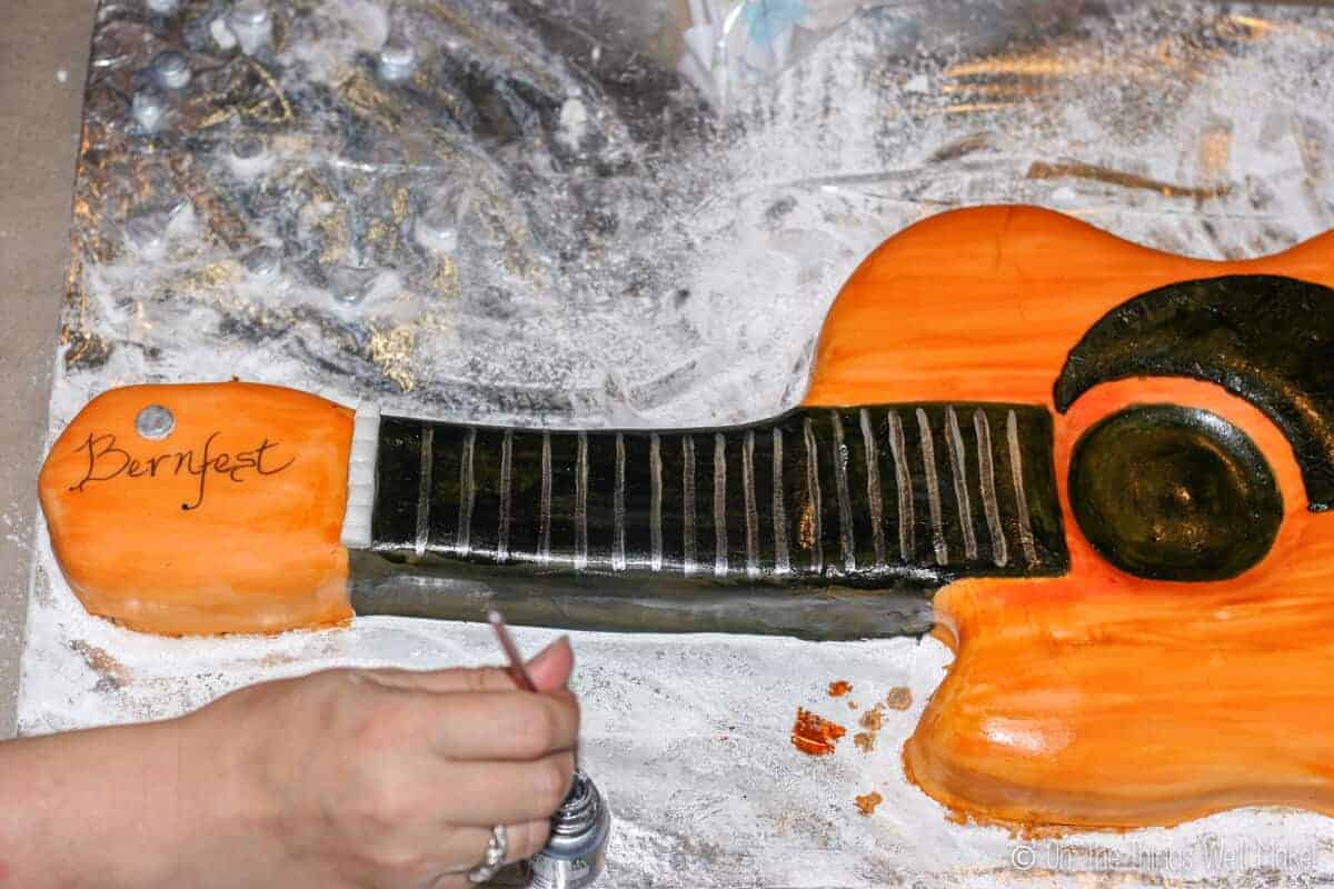 Top view of a hand with a paintbrush in food coloring ready to paint on more details of the fondant guitar cake.