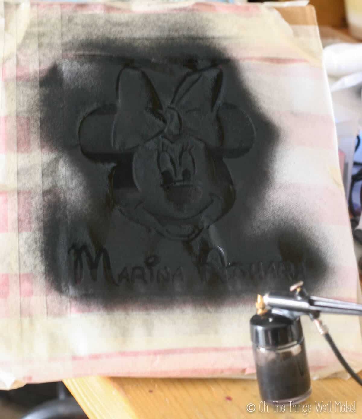 Black fabric paint being sprayed over a freezer paper stencil of Minnie Mouse with an airbrush.