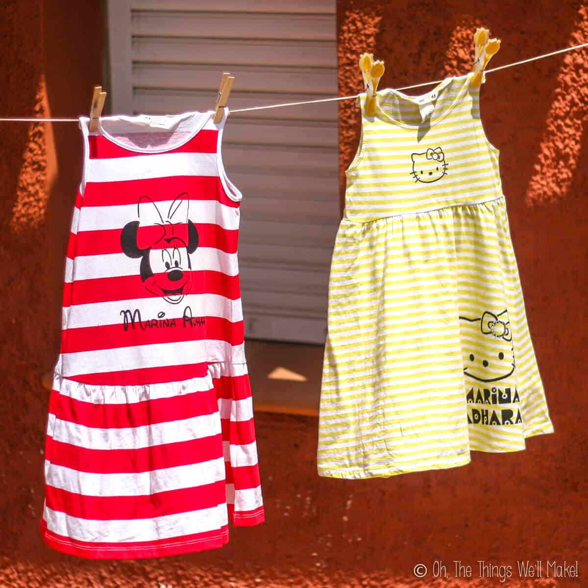 Two dresses hanging on a clothesline. A red and white striped sleeveless dress that has been painted with Minnie Mouse, and a yellow and white striped sleeveless dress painted with Hello Kitty.