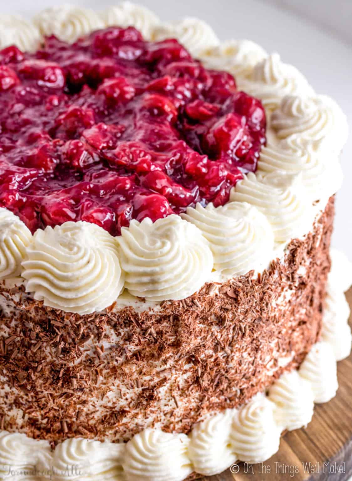 Overhead view of a black forest cake. Red cherries on top, white whipped frosting on the sides, and chocolate shavings on the side.