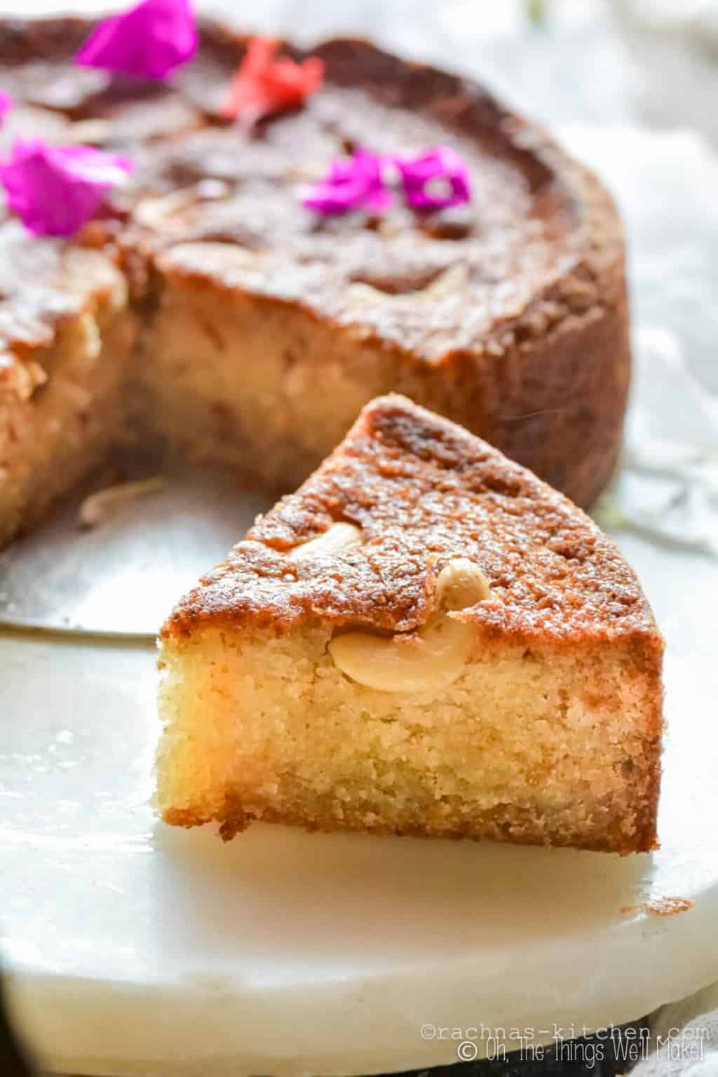 A slice of mawa cake in front of a full cake decorated with purple flowers.