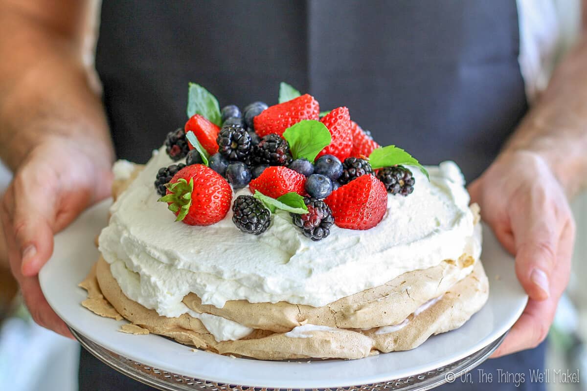 Hands holding a plate of pavlova. White icing on meringue decorated with red strawberries, blueberries, and black raspberries.