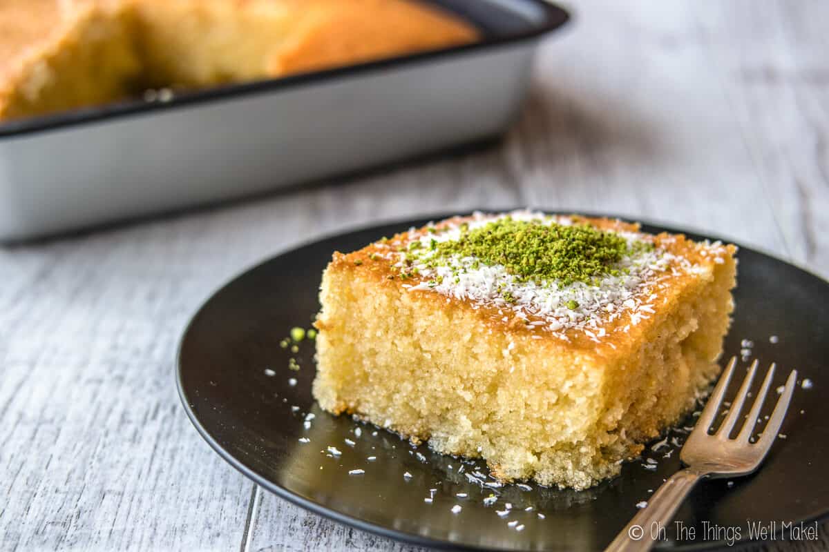 A slice of Greek Revani cake on a black plate, in front of a pan of it.