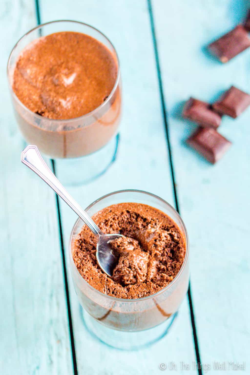 Overhead view of two glasses of chocolate mousse. Bottom glass has a silver spoon inside it and leaning to the side. A couple pieces of chocolate are laid on the side.