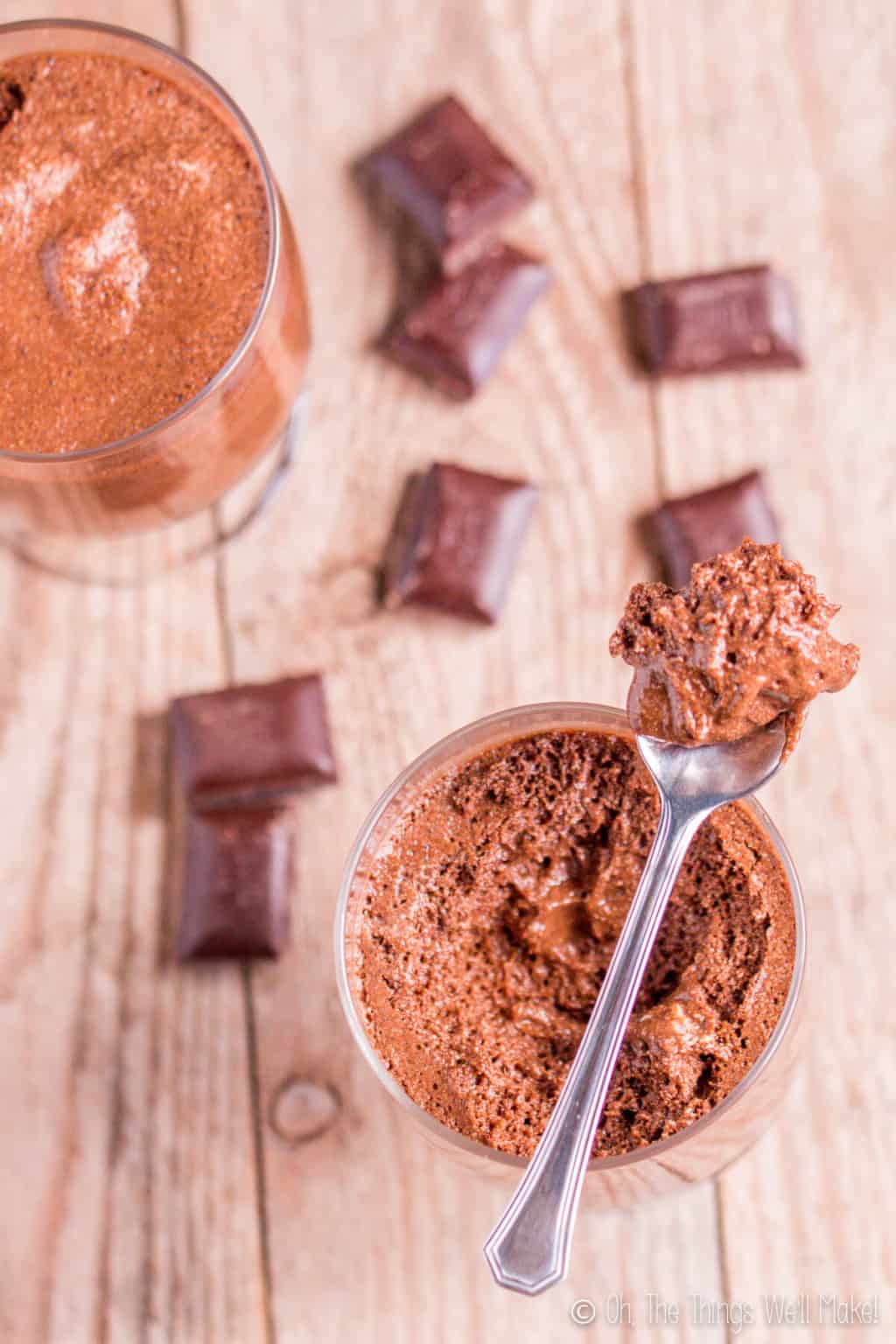 Overhead view of a glass full of homemade chocolate mousse, with a silver spoon on top full of some mousse. Pieces of chocolate surround the glass of chocolate mousse.