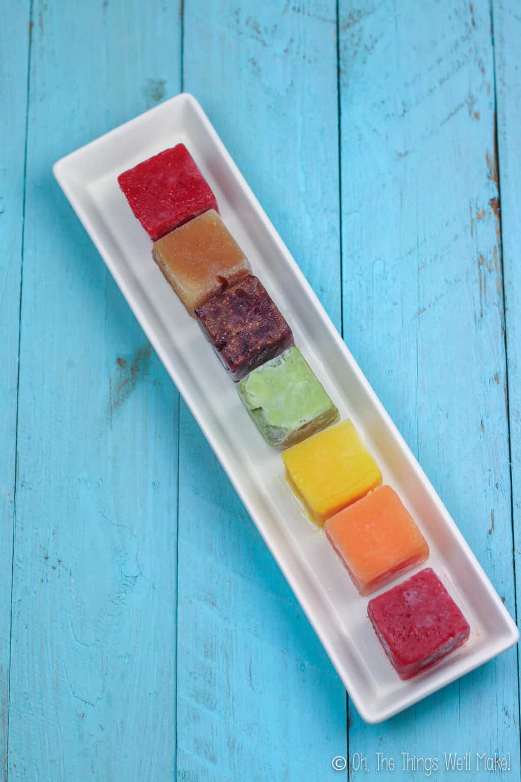 A rainbow of frozen ice cubes on a white rectangular plate over a blue wood surface.