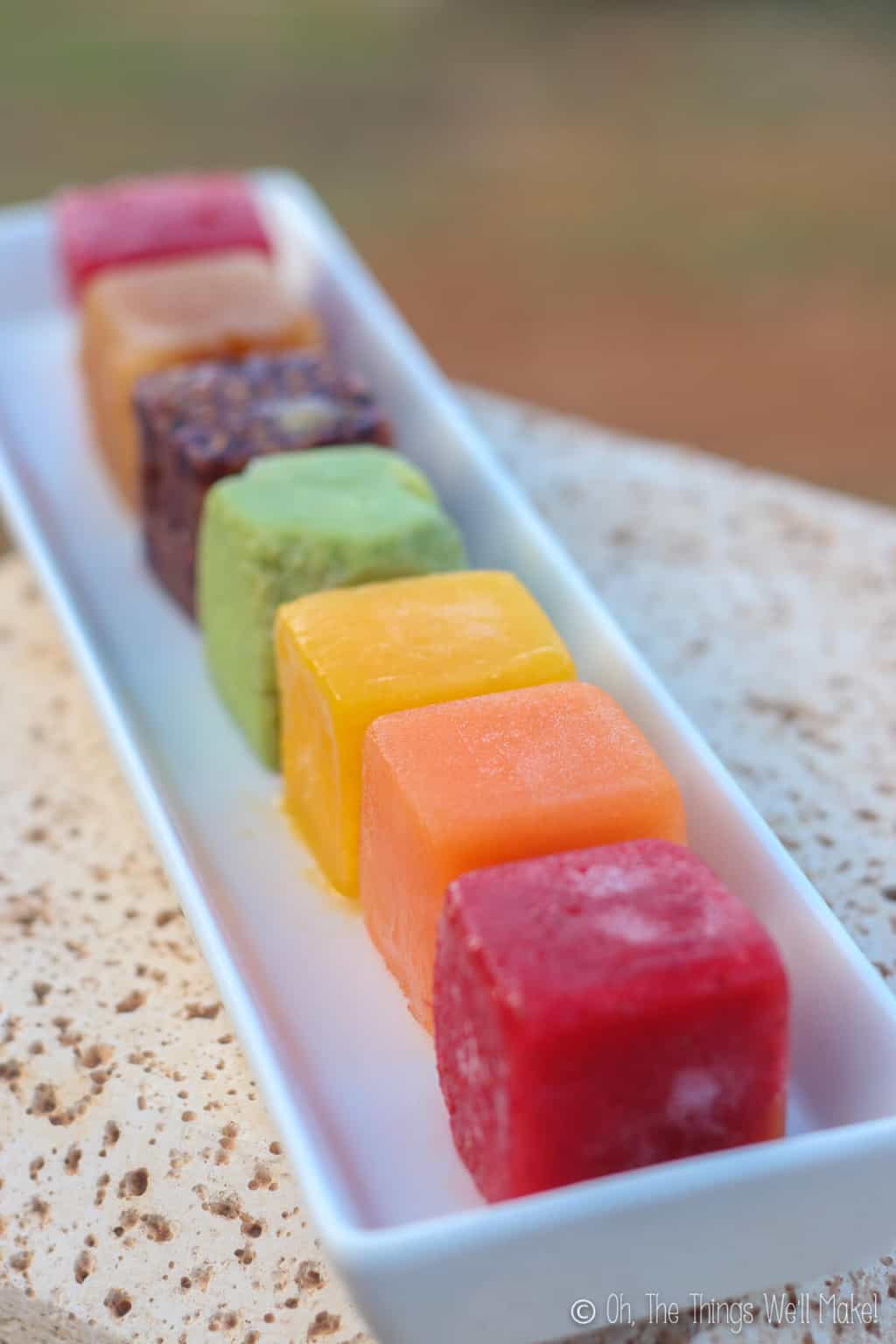 Close up of a rainbow of frozen ice cubes on a rectangular plate placed outside. The colors of the cubes, starting with the one in front is red, then orange, then yellow, then green, then purple, and then dark orange.