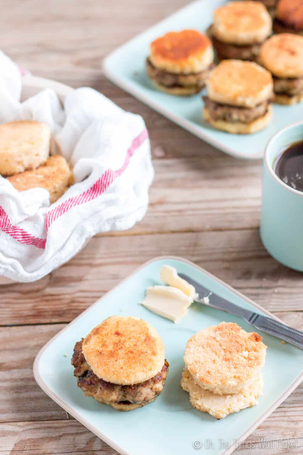 In the foreground, is a light blue square plate with one sausage biscuit and a couple of biscuit pieces next to a knife with butter. On the background is a bowl of biscuits and another plate of sausage biscuits.