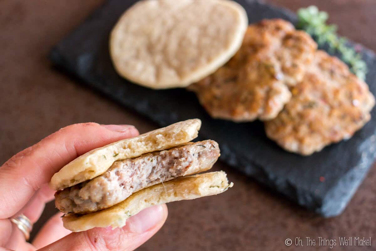 Close up of hand holding a homemade sausage patty between two paleo pitas with a plate of sausage patties and pitas in the background.