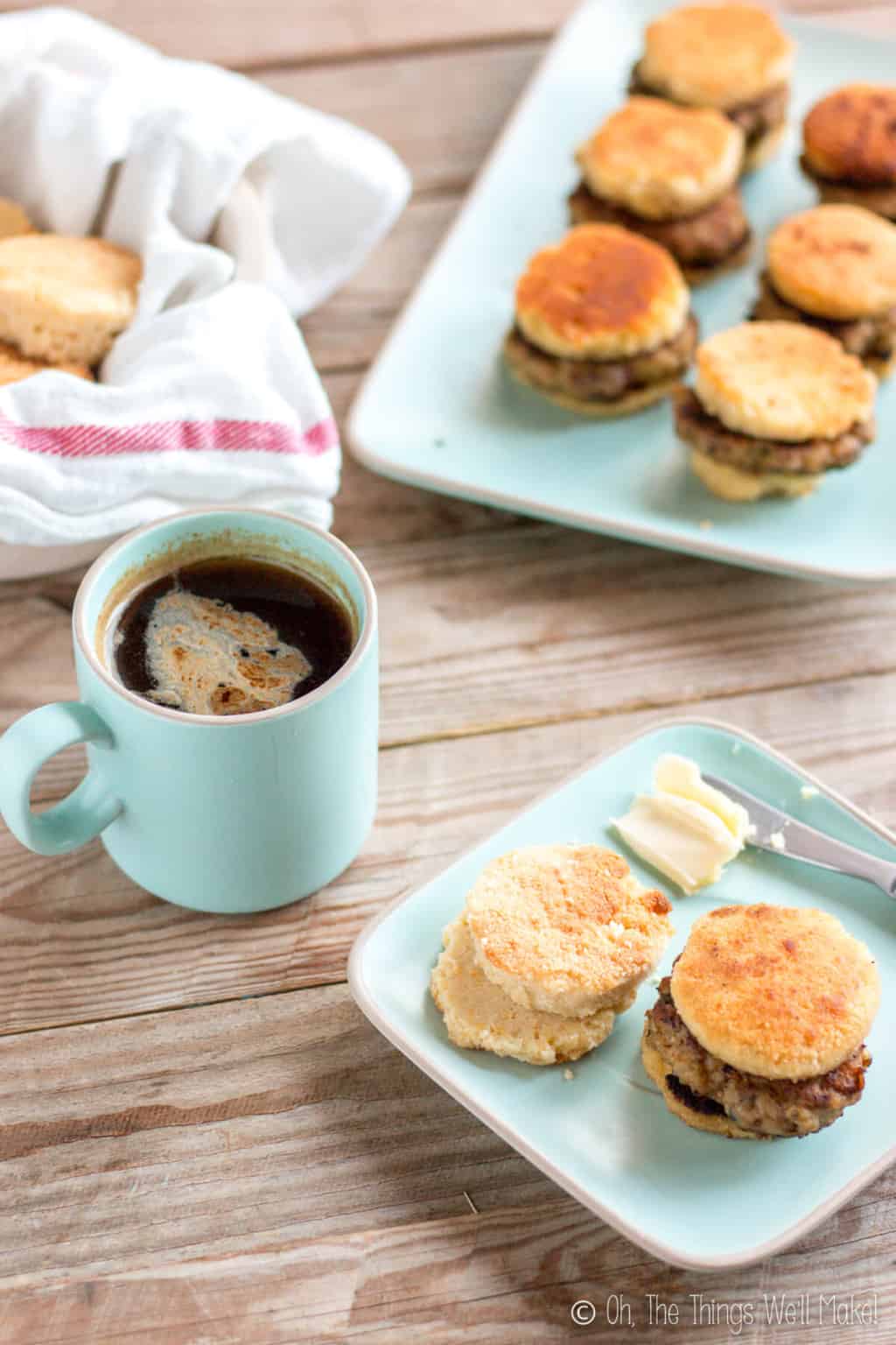 In the foreground, is a light blue square plate with one sausage biscuit and a couple of biscuit pieces next to a knife with butter. A cup of coffee is placed beside it with a bowl of biscuits and another plate of sausage biscuits in the background.v