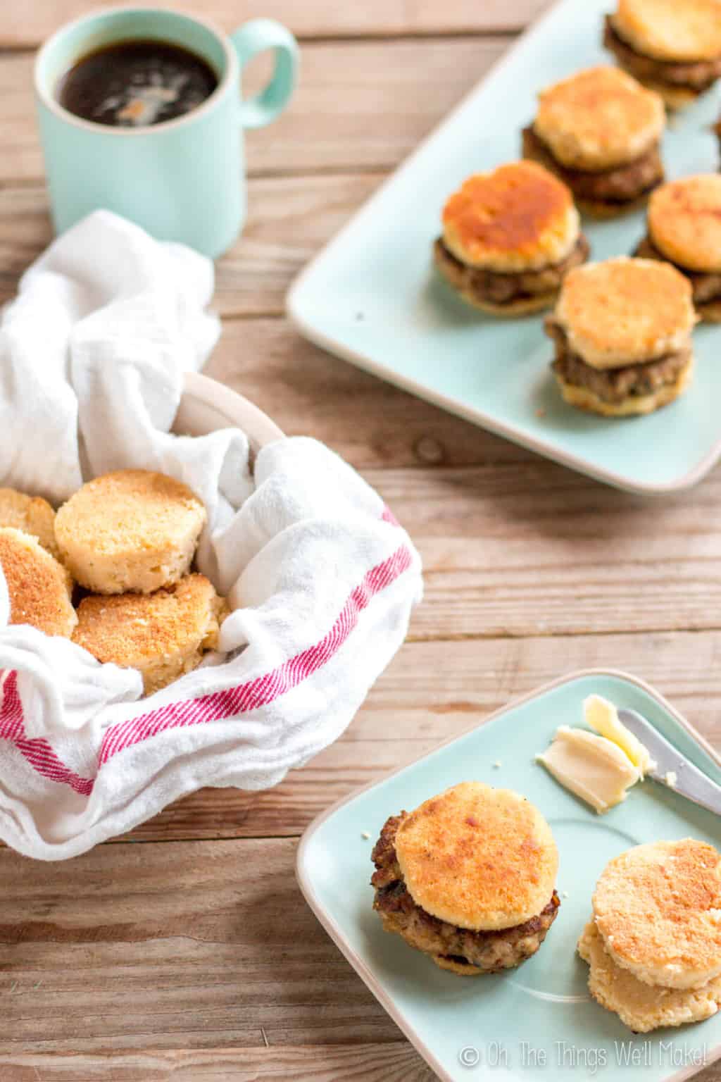 In the foreground, is a light blue square plate with one sausage biscuit and a couple of biscuit pieces next to a knife with butter. In the background is a bowl of biscuits and another plate of sausage biscuits.