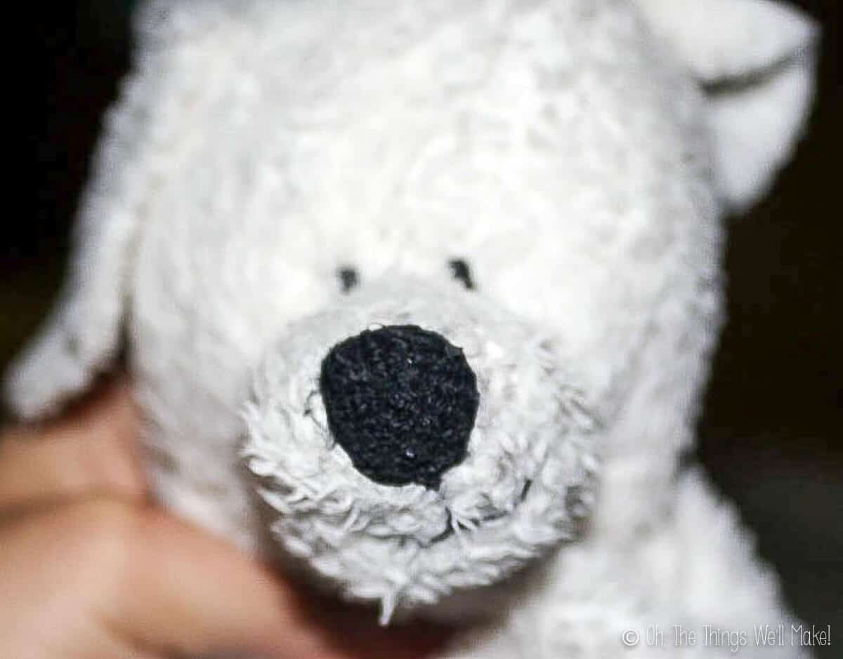 Close up of a hand holding the head of a white fluffy stuffed dog showing the black nose and eyes.