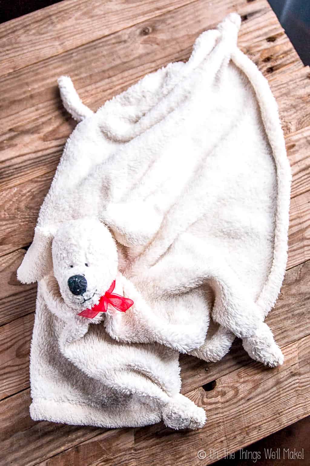 A white fluffy blanket with stuffed animal puppy head spread out on a wooden surface that shows off the head, tail, and corners of the blanket.