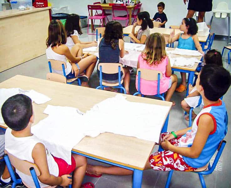 A group of children at tables with white t-shirts on them, ready for decorating.
