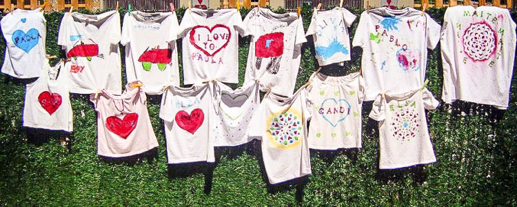 15 kids t-shirts that have been hand painted and hung on a fence.
