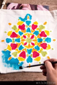 Painting on a paper snowflake stencil that has been adhered to a cloth bag, making a colorful, mandala-like design.