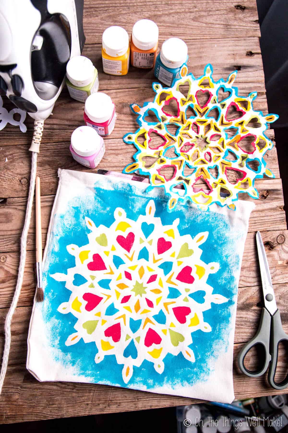 A bag with a hand-painted mandala design on it next to the stencil used to make it, fabric paints, a pair of scissors and an iron.