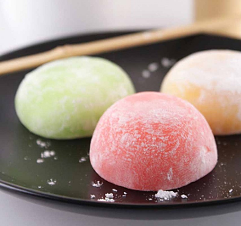 3 mochi on a black plate, one is pink, one is green, and one is a light yellow.
