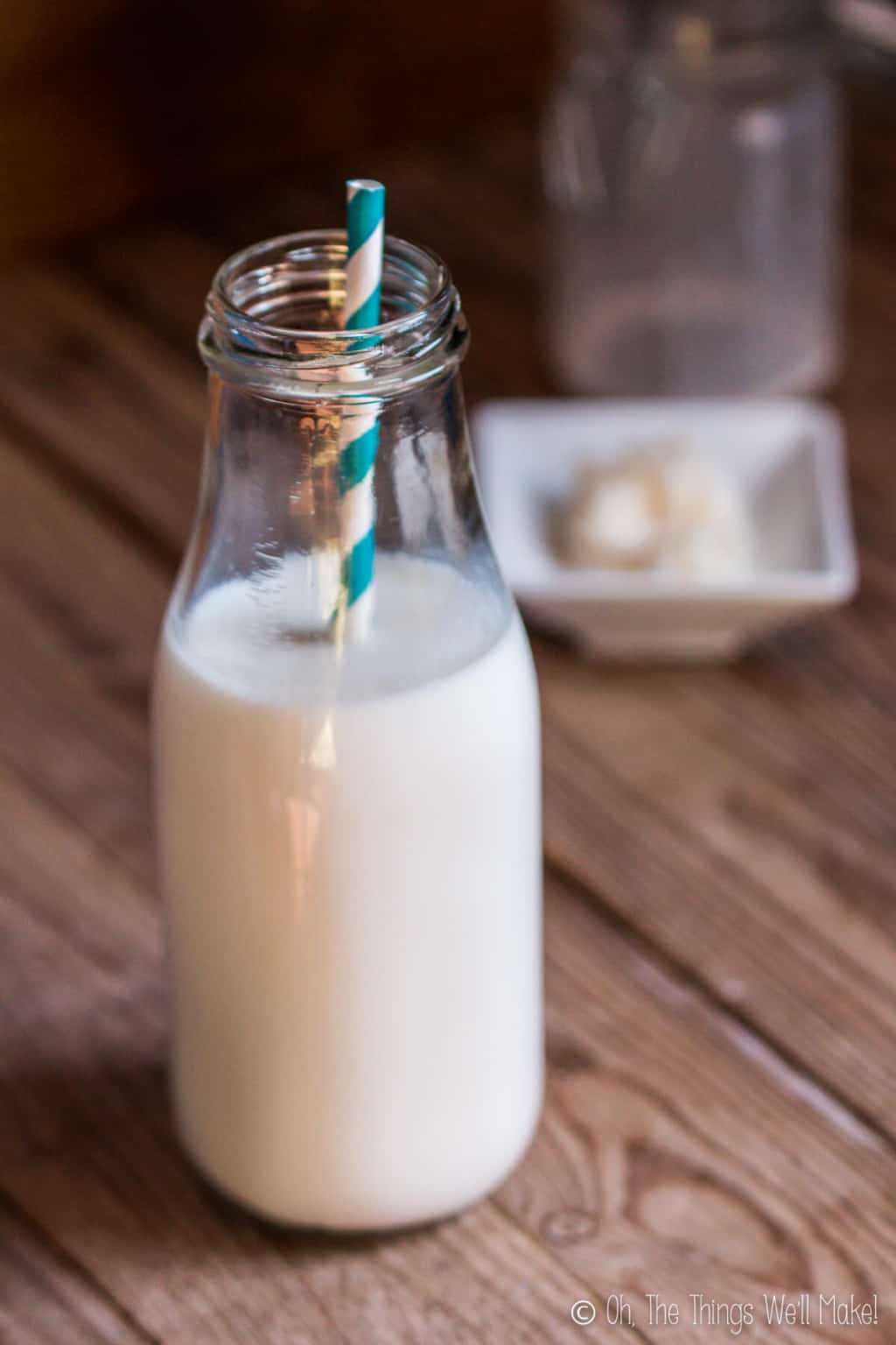 Close up of a bottle of kefir with a white and blue striped paper straw in it, next to  a small plate filled with some kefir grains.