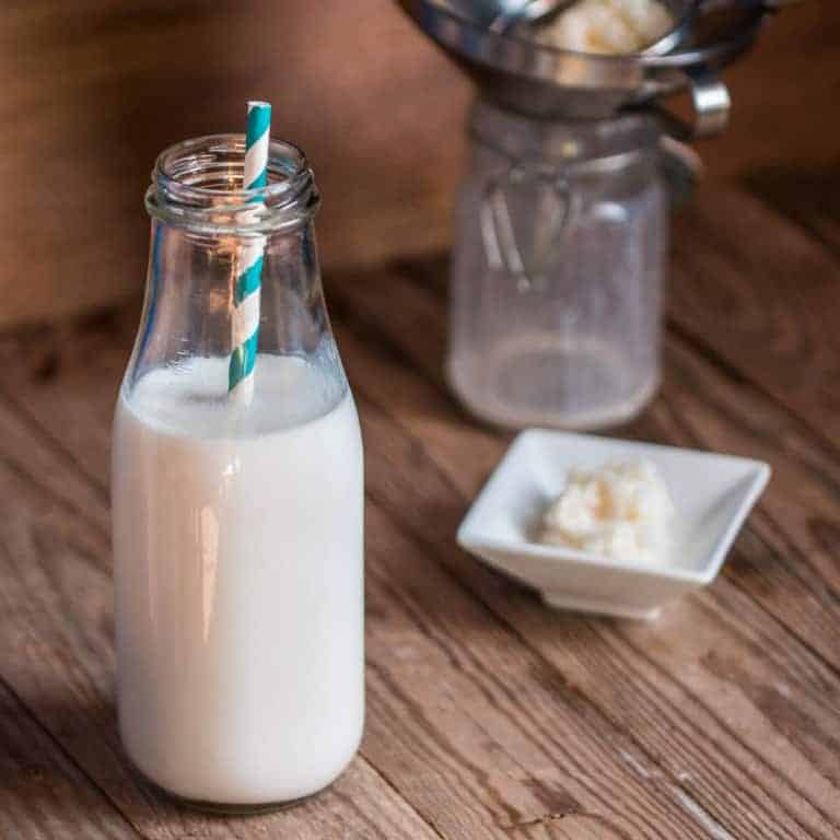 A clear glass bottle of milk kefir with a striped blue and white straw inside, and a small, white square plate of kefir grains in the back.