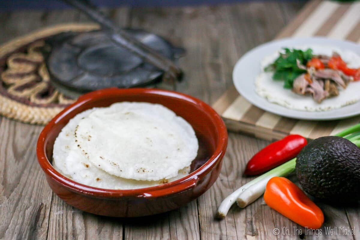 A terra cotta pan filled with homemade corn tortillas on the left side with some vegetables on the other side and a plate of garnished pork carnitas on a tortilla.