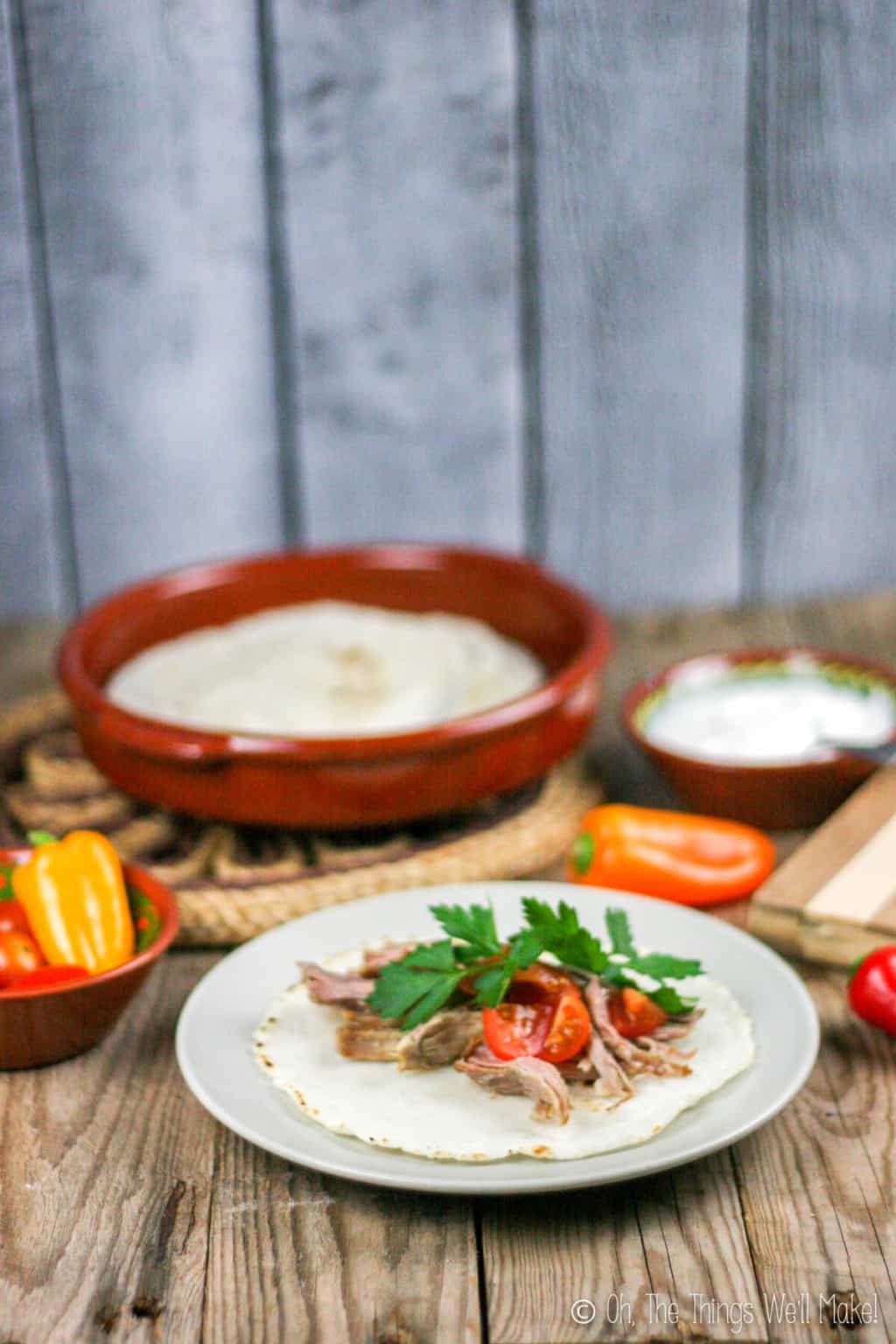 Pork carnitas on a corn tortilla on a plate, garnished with parsley and tomatoes. In the background, more tortillas can be seen. 