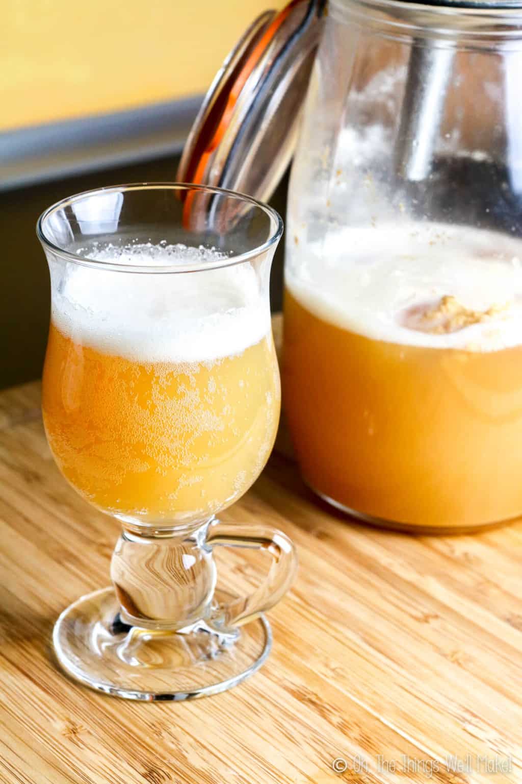 A glass full of homemade kefir hard cider beside a large jar full of the same cider on a wood surface.