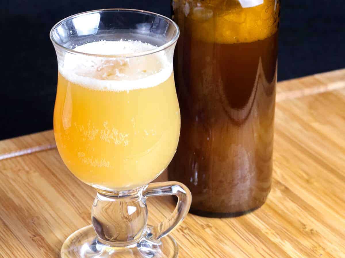 A glass full of yellow homemade hard cider made from kefir grains, next to to a bottle full of the same cider.