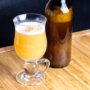 A glass full of yellow homemade hard cider made from kefir grains, next to to a bottle full of the same cider.