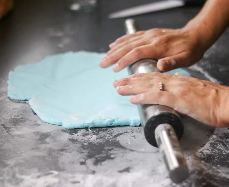 hands rolling out blue fondant with a rolling pin.