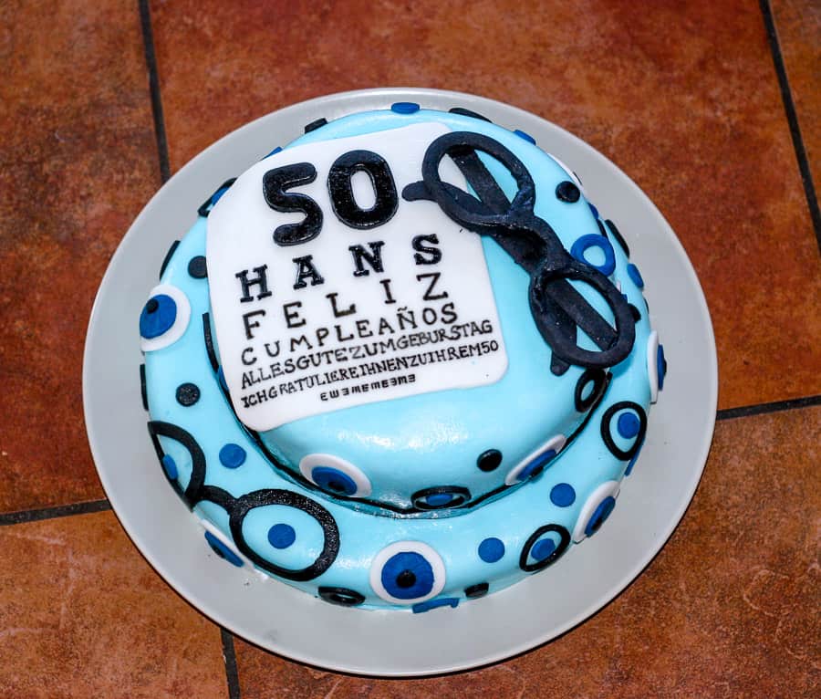 Top view of a blue covered fondant birthday cake decorated with eyes, glasses, and an eye chart with a birthday message in fondant saying “50 Hans Feliz Cumpleaños”