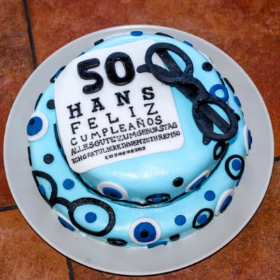 Overhead view of a fifty year old birthday cake covered in light blue fondant and decorated with fondant decorations of black eyeglasses, eyes, and a birthday greeting that says "50 hans feliz cumpleanos"