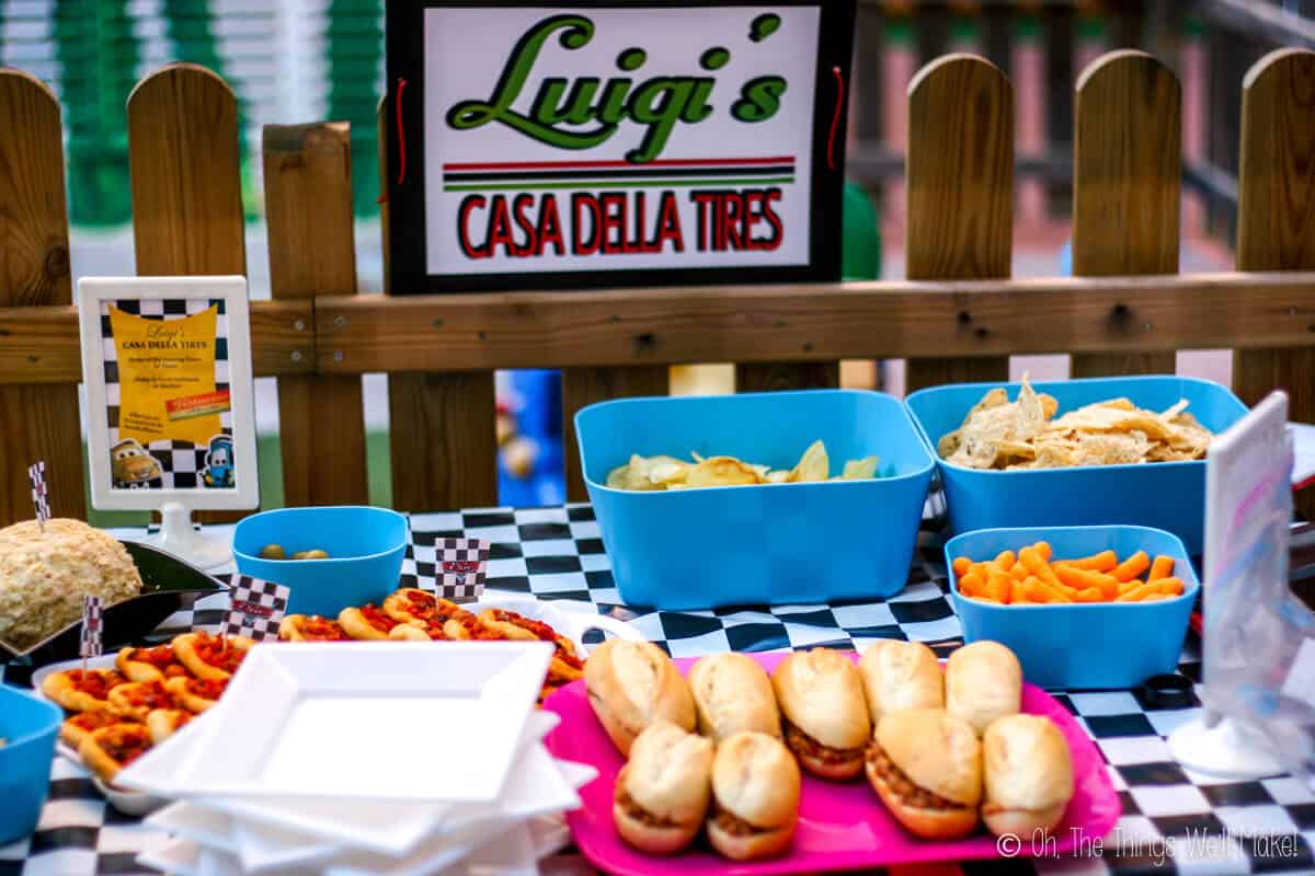 Sandwiches laid on a pink tray with other party snack foods on a table covered in checkered tablecloth, with a sign on top that says "Luigi's Casa Della Tires"