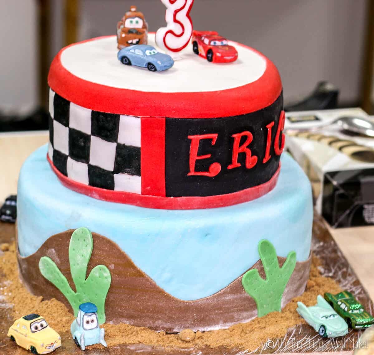 Two tier Cars themed fondant cake decorated with a painted checkerboard  pattern in the top layer and cactus with desert hills in the bottom layer, with several fondant car figures laid around.