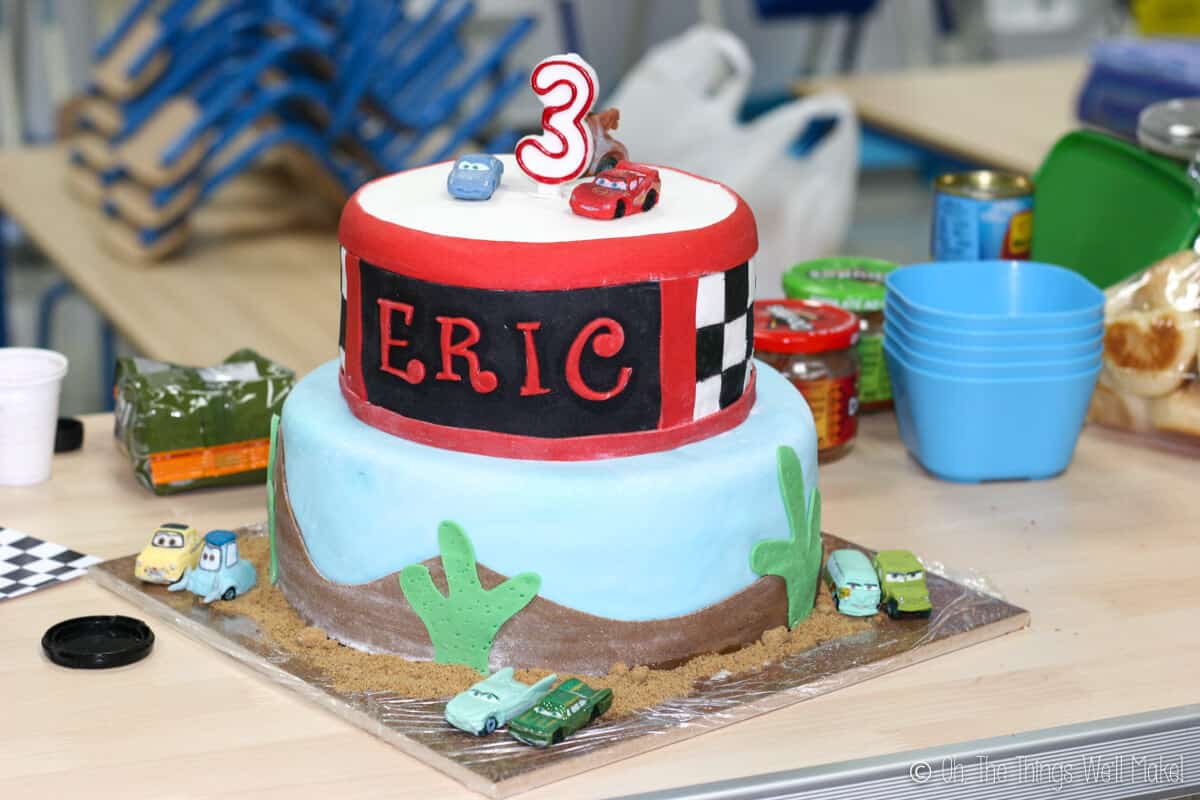 Close up of a 2 tier Cars themed fondant cake with fondant cars all around the cake. Bottom layer is light blue decorated with brown fondant as sand and green cactus shaped fondant. Top later is red with checkered flag and a black space with the name Eric decorated on it. Fondant cake topped with a number three candle, Lightning McQueen, Mater, and Sally fondant decor.