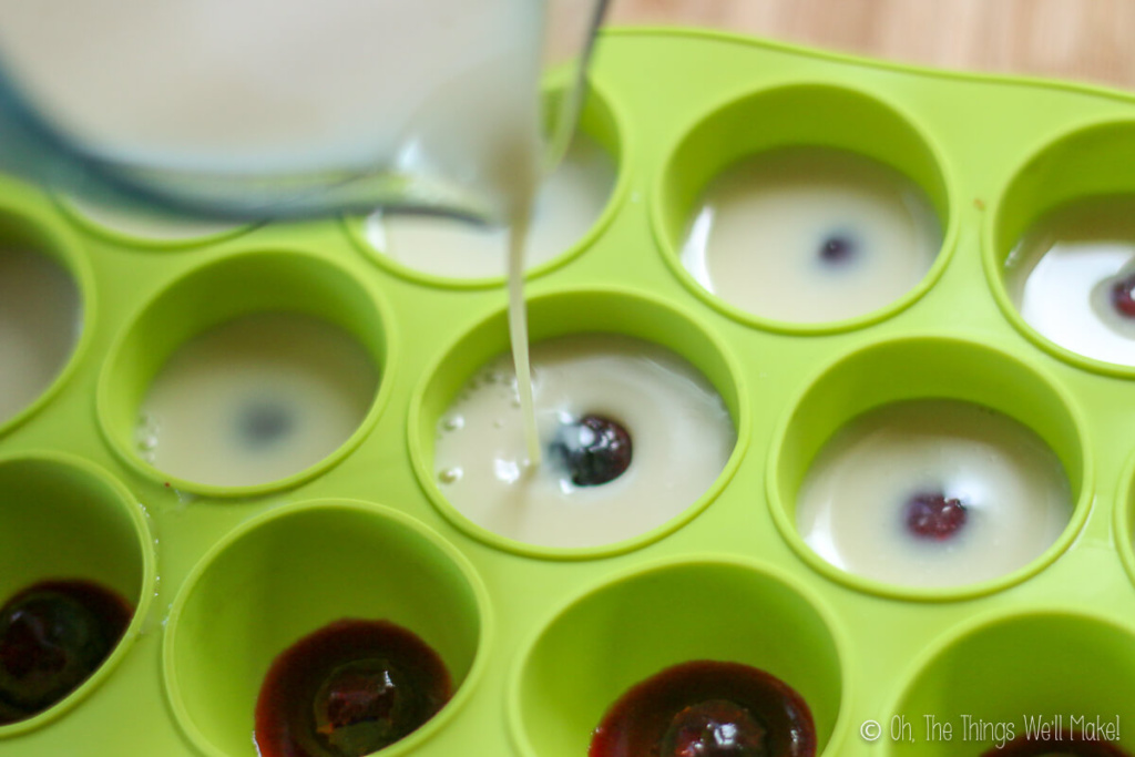 Pouring a white gelatin mixture over blueberries and red gelatin in silicone ice cube trays.