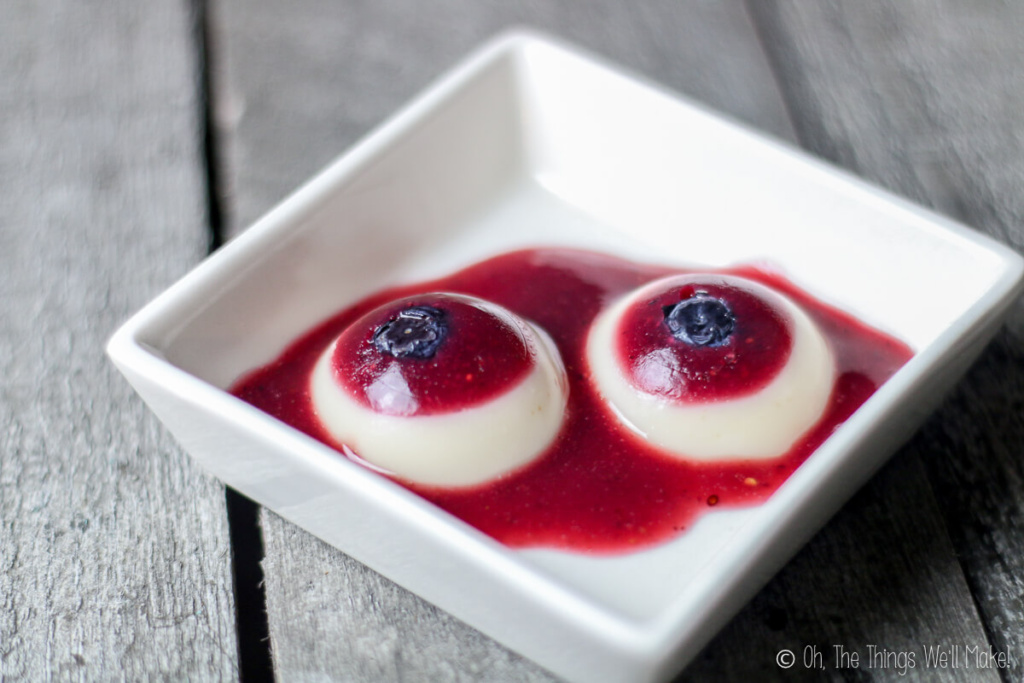 Two gummy eyeballs in a square bowl served with berry puree, placed on a grey wood surface.