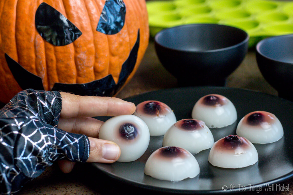 A hand holding a gummy eyeball from a plate full of gummy eyeballs and a jack-o-lantern in the background.