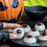 A hand holding a gummy eyeball from a plate full of gummy eyeballs and a jack-o-lantern in the background.